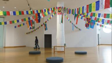 A large pillar stands in the middle of a gallery wrapped in white cloth, with several strings of colourful bunting carrying the flags of various countries and Tibetan art tied to the top of the pillar where a red and yellow flag has been pinned. There is a small wooden table at the base of the pillar as well as black circular cushions placed around it. Two people dressed in jeans and dark T-shirts are walking across the gallery