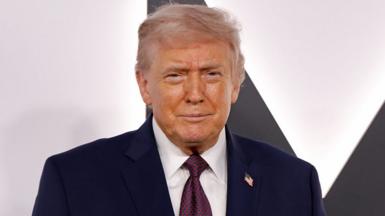 Donald Trump in a blue suit and blue tie with red dots stands in front of a white background