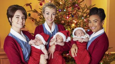 Three actors from Call the Midwife hold babies, dressed in Santa outfits, with a Christmas tree behind them 