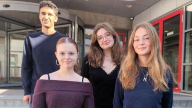 Four young people stand on steps outside their school - one male and three females - looking at the camera with faint smiles.