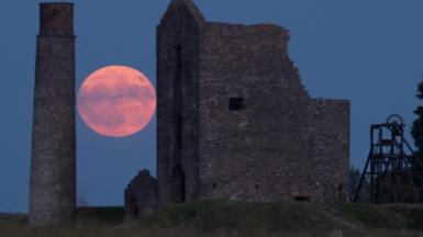 Orange and purple hues of Strawberry Moon positioned between ruins of a building and an old monument illuminating a blue night sky