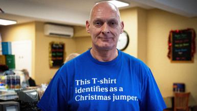 A bald man in a royal blue T-shirt bearing the words "This T-shirt identifies as a Christmas jumper" stands with his back to the counter in his coffee shop, a customer on the other side of it.