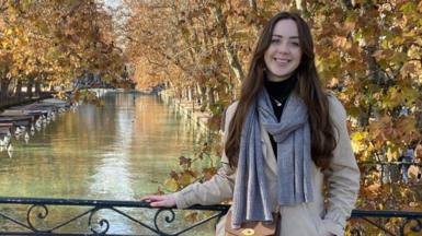 Darcie James standing on a bridge overlooking a canal in France, on her year abroad. She has long brown hair and is wearing a coat and scarf and smiling at the camera.