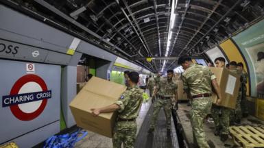 Soldiers in combat gear move boxes off pallets on a disused Tube platform. 