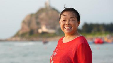 A smiling Su Min in a red top standing by a water body with a blurred hill in the background.