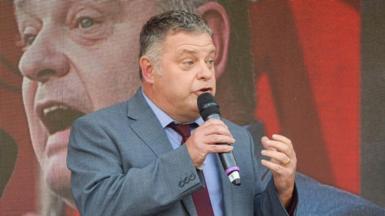 Mike Amesbury speaking into a microphone on stage at an outdoor meeting in fron of a big screen