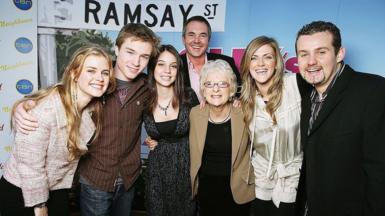 Seven people stand with their arms around each other's shoulders in front of a partition with the graphic of a street sign saying "Ramsay St"