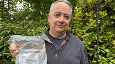 A man in a grey quarter-zip holding a crumpled package from Royal Mail with an apology letter, stood in front of a green bush.