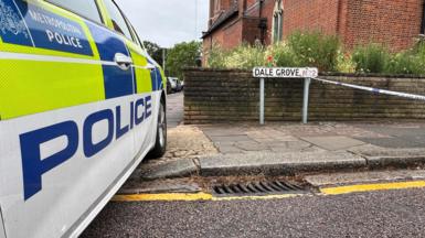 Metropolitan Police car parked beside a street sign reading "Dale Grove N12", with police cordon tape strung across the road and a red-brick church behind.
