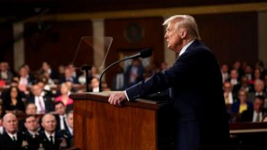 Trump is stood at a podium facing to the left of the image, speaking to a crowd of faces in the House of Representatives