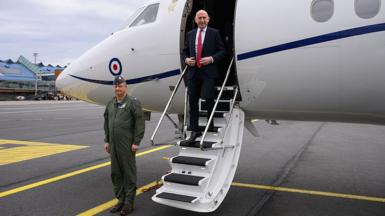John Healey in a white shirt, black suit and red tie, seen exiting an RAF plane at an airport as a soldier stands to attention nearby