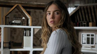 Sydney Sweeney appears with her head turned to face away from the camera. She is wearing a white long-sleeved ribbed top and is in front of a dolls house.