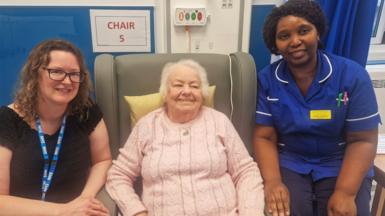 Picture of 86-year-old Shirley Xerxes from St Albans who was one of the first people in the UK to be given the new Keytruda injection. She is smiling and sitting on a chair in a hospital clinic wearing a light pink jumper. So her left and right are members of NHS staff.
