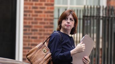 Lucy Powell is holding a brown folder and has a large brown-coloured bag over her right shoulder. She is wearing a dark blue jacket. A brick wall and a metal fence can be seen in the background.