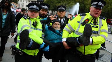 A male protester wearing a hat and making a peace gesture with his fingers is carried away by four police officers