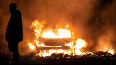 The silhouettes of a man shrouded in darkness with a large fire coming from a burning car in the background.