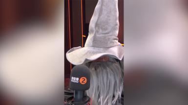 Scott Mills sits in front of a microphone. He wears a large grey wizard's hat and a long grey fake beard