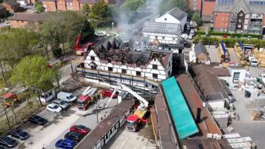 Aerial view of a large Tudor‑style building severely damaged by fire. Most of the roof has collapsed, leaving charred beams exposed and smoke rising from the structure. Firefighters use hoses and an aerial ladder platform to extinguish remaining hotspots, with several fire engines positioned around the scene. 