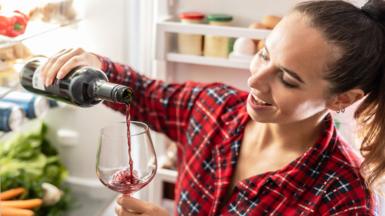 Wine bottle tilted over a wine glass as red wine is poured into the glass in the foreground. A person wearing a red checked shirt holds the bottle and glass. An open refrigerator is visible in the background, with shelves containing eggs, containers and vegetables.
