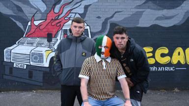 Three men pose in front of a mural showing a burning PSNI vehicle. The man in the middle is wearing a tri-colour balaclava. The man on the left is wearing a grey jacket and the man on the right is wearing a black jacket.