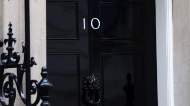 A black door in Downing Street with the Number 10 on it. A black railing can be seen in the foreground.