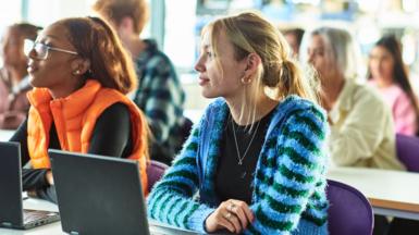 University students sitting in a lecture. The two women in the front row are wearing an orange gillet jacket and a fluffy stripey blue and green cardigan.