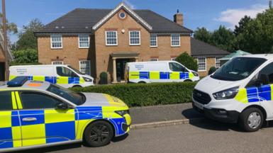 A police car and a few forensic vans are parked outside a large house, some on the street, some on the driveway