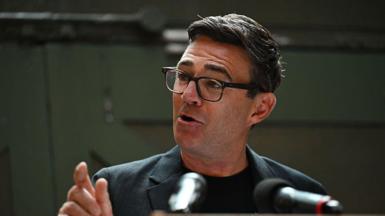 Andy Burnham making a speech at the People's History Museum in Manchester on Monday. He is gesturing with his right hand and is mid-speech. He is wearing glasses, a grey blazer and black shirt.
