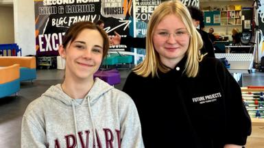Two girls smiling at the camera as they pose for a picture in a youth club. Maisie on the left has brown hair pulled back in a pony tail and is wearing a grey hoodie with Harvard University branding in large red type. Iga on the right wears a black hoodie bunched up on her arms. She has shoulder-length blond hair and has rimless glasses.