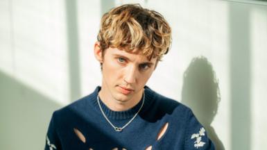 A portrait of Troye Sivan where he is looking at the camera with brownish hair. He has a nose ring on, a blue jumper and a necklace. He's infront of a white wall. 