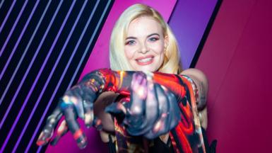 A woman with blonde hair wearing a multicoloured dress points at the camera while smiling in front of a pink neon backdrop.