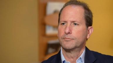 Headshot of Jon Brilliant who is looking to the side of the camera. He has short dark hair and faint beard stubble. He is wearing a blue open-necked shirt and navy suit jacket. He is pictured in a room with painted yellow walls and a wooden bookshelf behind him. 