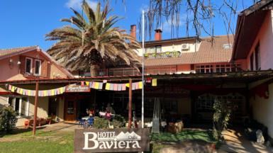A sign reading "Hotel Baviera" stands in front of a building with a red-tiled roof. Bunting hangs over the entrance. A palm tree towers over the building.
