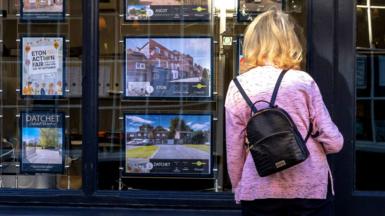 A woman wearing a pink jacket and a small black rucksack looks at adverts in an estate agent's window