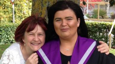 Jill and June after a graduation ceremony. Jill, on the left, is wearing a white lace top and June is wearing a black and purple graduation gown. They are standing in front of a tree.