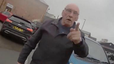 A man standing in a car park. He has his finger pointed at the person wearing the body-cam.