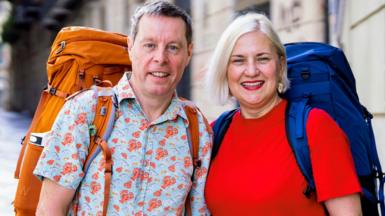 Mark Blythen, in his 60s, who has short grey hair and is wearing a short sleeved blue shirt with a lobster pattern and a large orange backpack, stands next to Margo Oakley, who has short blonde hair, a red t-shirt and a large blue backpack. 