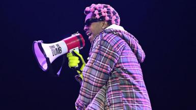 A$AP Rocky performing on stage. He wears a checked jacket and has pink rollers in his hair. He is holding a microphone with 'Don't Be Dumb' on it.