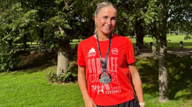 Leah smiling looking at the camera. She is stood in front of trees and greenery and wears a red Cardiff half marathon finishers t-shirt, a black medal and black running shorts. Leah has blonde hair in a high ponytail. 