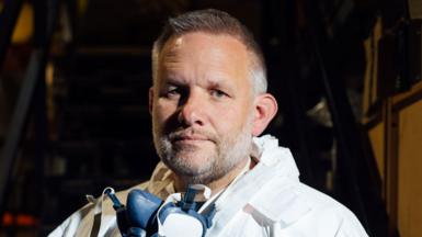 Up close photograph of Ben Giles. His is aged 49, has grey short hair. He wears a  face mask around his neck. You can see his PPE around his neck, which is a hooded white suit. 