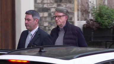Lord Mandelson being arrested at his London home on Monday - He has dark grey hair swept back, is wearing glasses, and has a serious look on his face as he walks away from his house