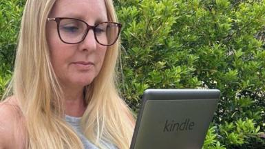 Kay, wearing a grey vest top and glasses and stood in front of a green bush, holds her 5th Generation Kindle in front of her