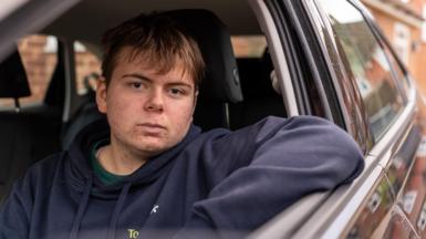 Young 18 year old man sits in the passenger seat of a black VW car, his arm resting on the open window as he looks to camera