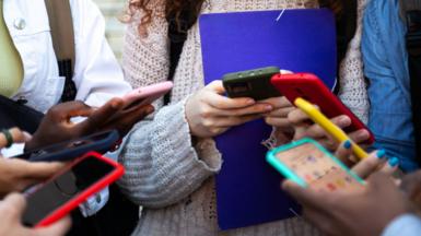 A group of children stand in a circle, all of whom are holding smartphones in brightly coloured cases. Their faces are not visible.