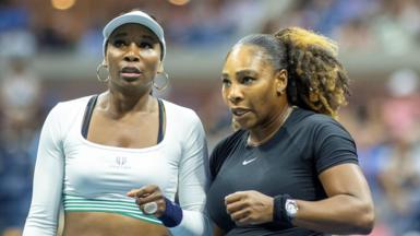 Venus Williams, in short white top and cap, stands alongside Serena Williams, who wears a black outfit