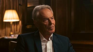 Sir Tony Blair sitting in a dimly-lit room, wearing no tie with his shirt's top button undone. He's looking to the side, off-camera. A lamp is lit behind him on the table