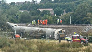 Three derailed train carriages lie at the bottom of a railway embankment near Lewes. Emergency vehicles, fencing and workers in high-visibility clothing can be seen around the site, while crews work on the railway line above.