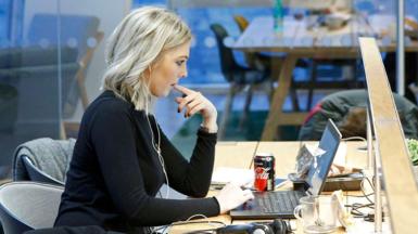 Woman with blonde hair sitting at a desk in front of a laptop in a well- lit office. She is wearing a black top and a pair of white in-ear headphones in her ears. She is holding her left index finger in her mouth while using the trackpad on the laptop keyboard. 