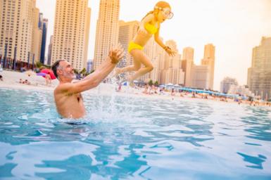 A man in the water at the beach in Dubai throwing his young daughter in the air. There are skyscrapers in the background