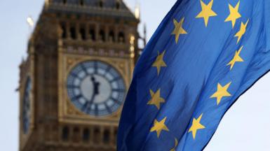 An EU flag blowing in the wind with the clock face of Big Ben visible in the background. The weather is sunny and the sky is light blue. 